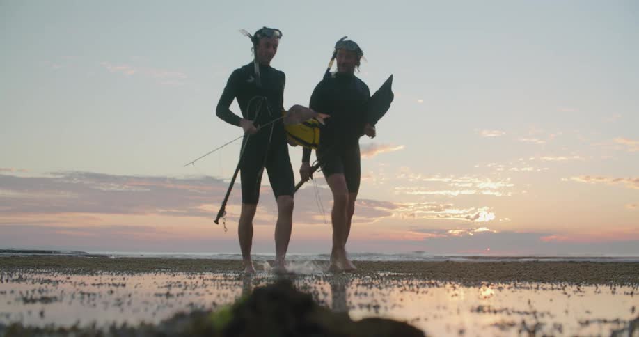 Two men with scuba gear walking on a beach with the sky and clouds in the background