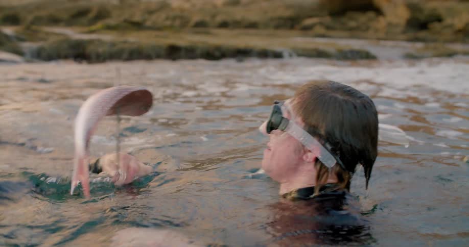 Two snorkelers rise up from the ocean and one of them shows off the fish he caught on a spear