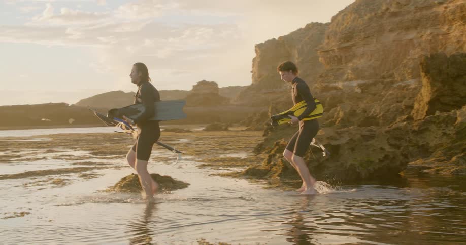 Two men wade into the ocean with their arms full of scuba gear