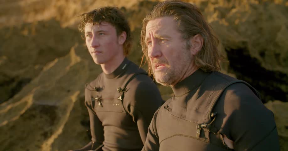 A father and son in scuba suits sit on rocks along the shore and talk