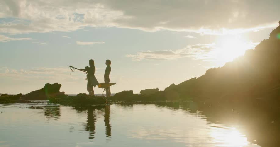 The silhouette of two men in wet suits carrying flippers and a spear walking on rocks with shimmering water and glaring sun