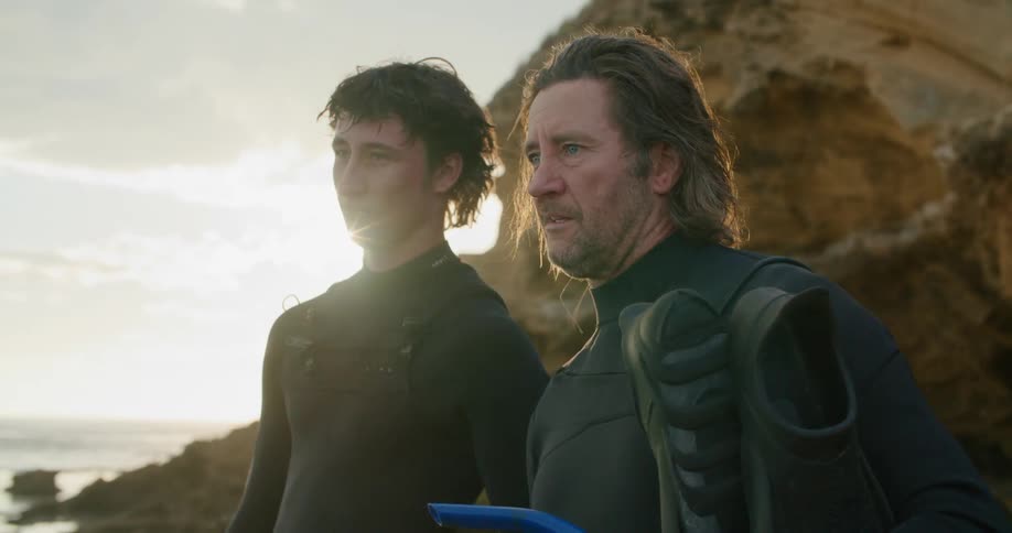 A father and son stand along a rocky coastline with spear fishing equipment while discussing their plan