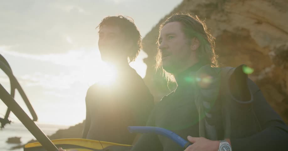 A man talks and points with a spear gun as his friend laughs while they both stand on shore wearing wet suits and carrying flippers