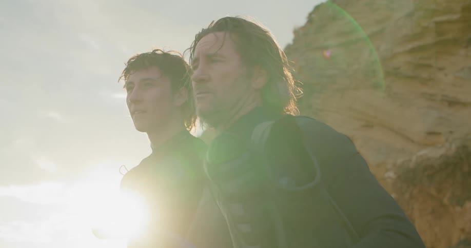 Two men wearing surfing suits standing in front of a flare of sunlight next to a cliff side having a conversation