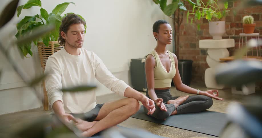 A man and woman meditate with their legs crossed while sitting on yoga mats in their living room