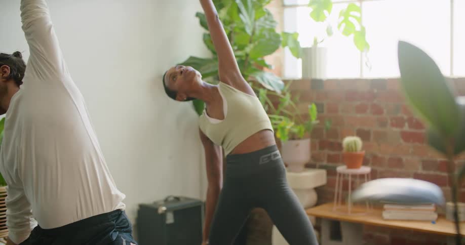 A man and woman stretch and lean forward while doing yoga in a room full of plants