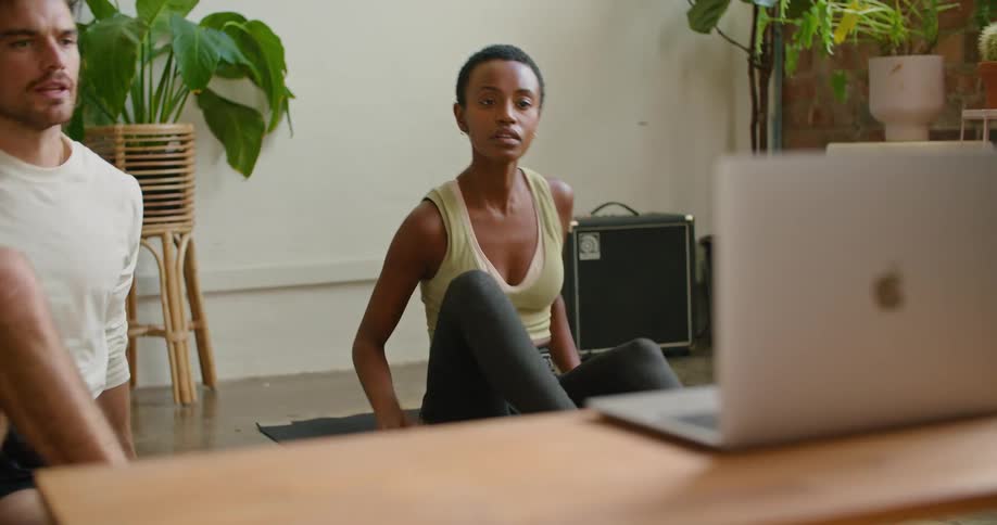 A man and woman sit on the floor while stretching as they watch the screen of a laptop