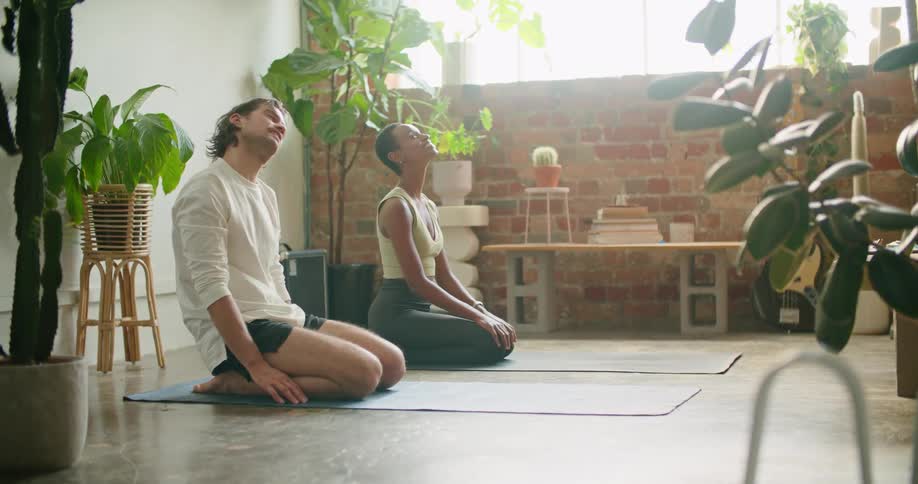 A man and a woman in a room on the floor stretching with plants and a wall in the background