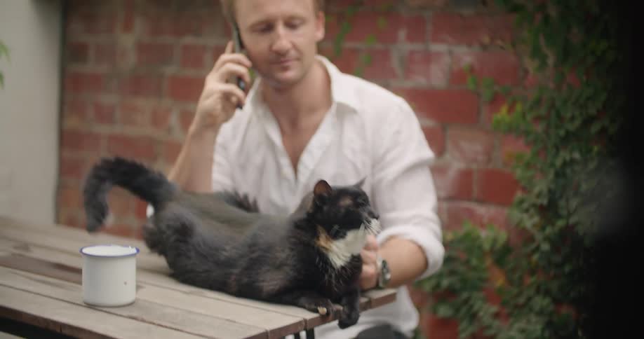 A man sits outside talking on the phone and petting a cat sitting on a table