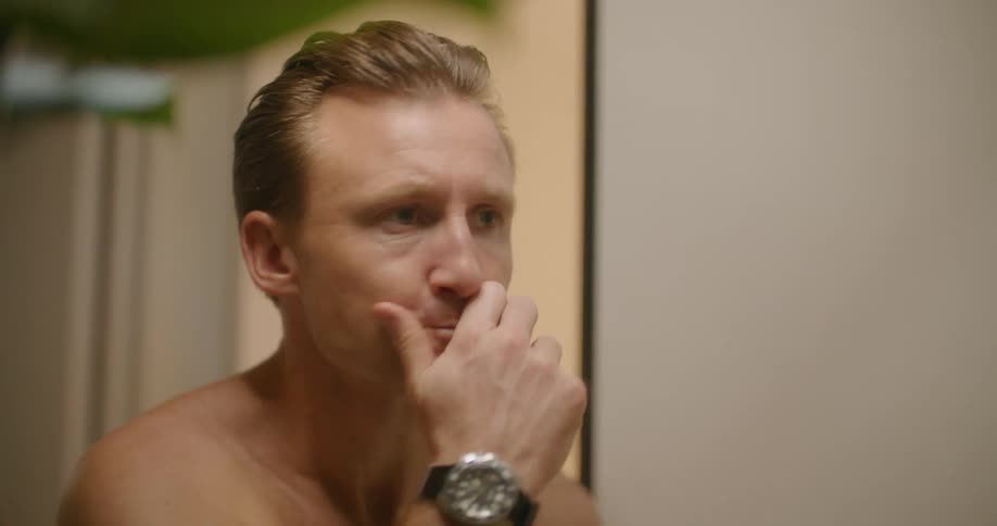 A shirtless man smiles at himself in the mirror after brushing his teeth