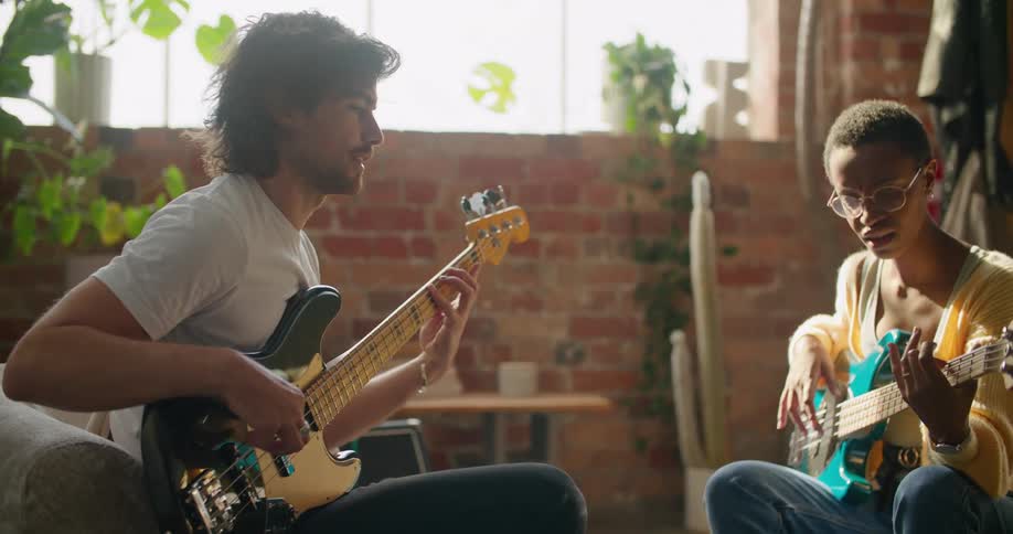 A guitarist attempts to teach a young woman how to play the guitar as they sit together in the living room of a house during the day
