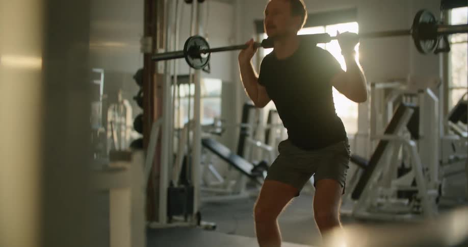 A man in the gym squatting with weights with equipment and windows in the background