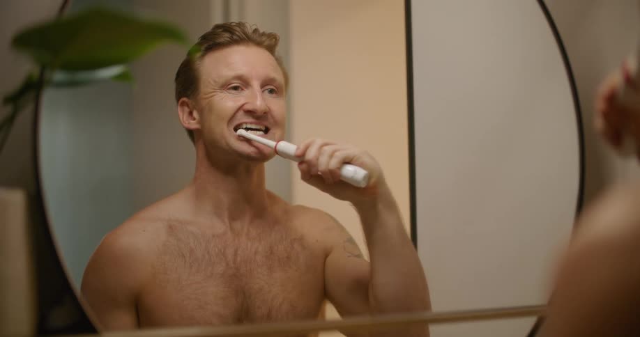A man brushes his teeth with an electric toothbrush in a round mirror