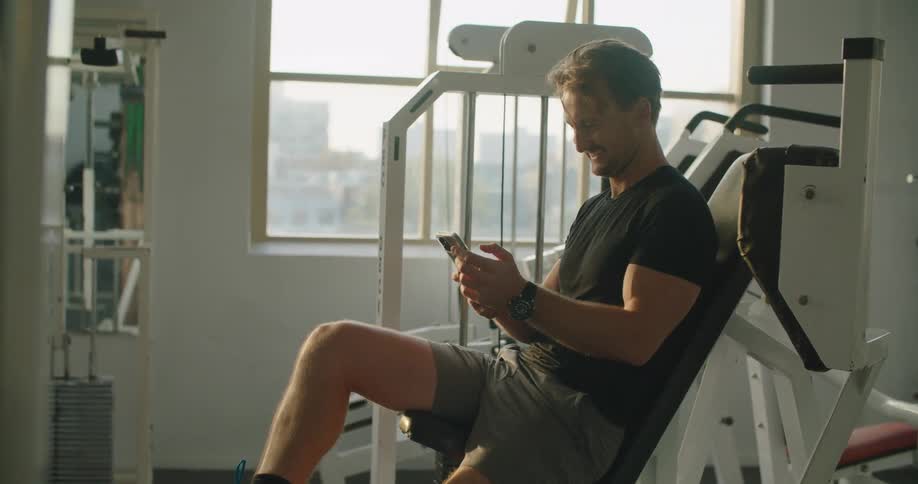 A man holds his smart phone in his hands while taking a break while in a fitness room