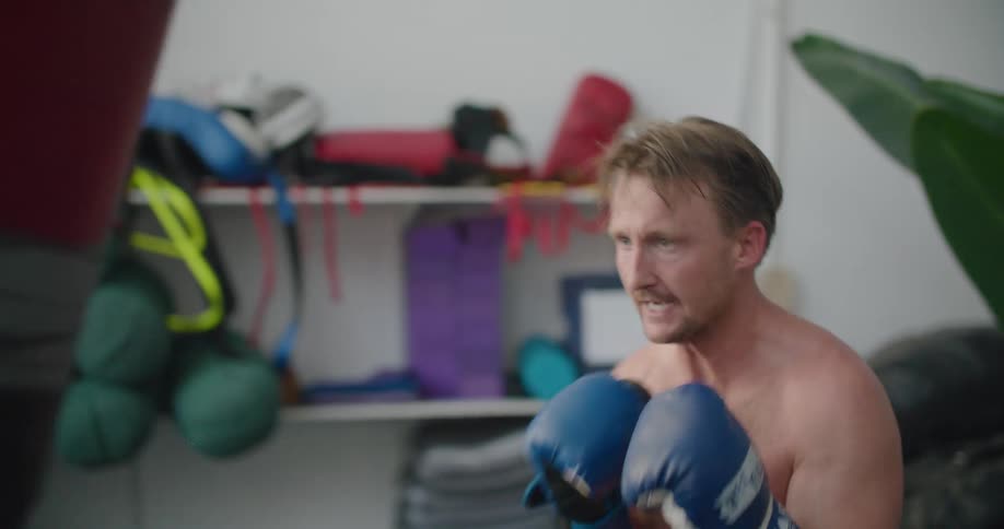 A shirtless man wearing boxing gloves repeatedly punches a heavy punching bag