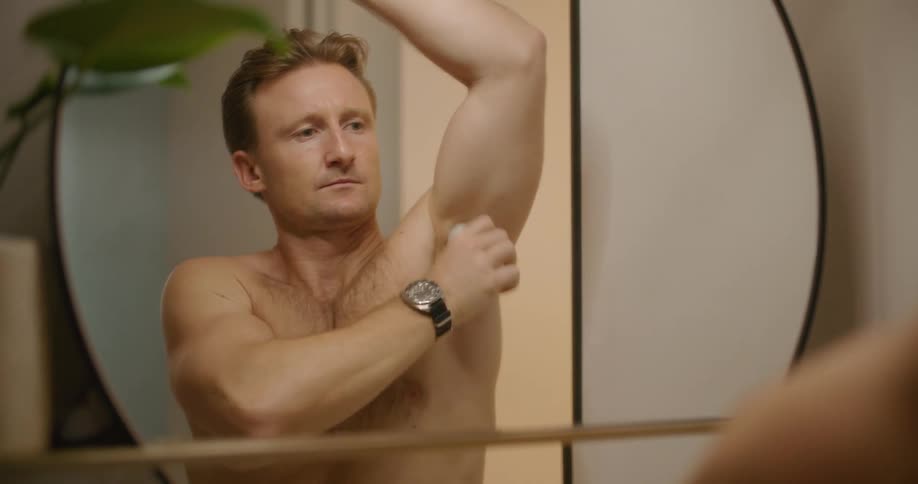 A man applies deodorant to his underarms in the bathroom mirror