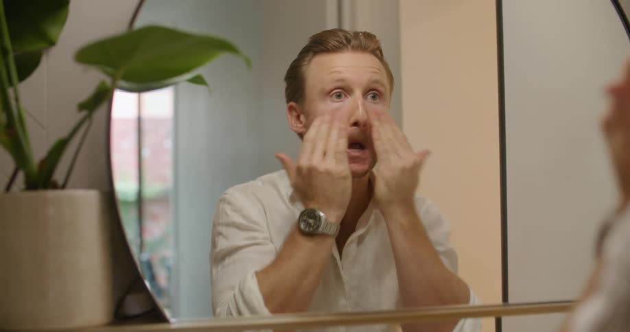 A man looking in the mirror rubs moisturizer on his face as he is getting ready