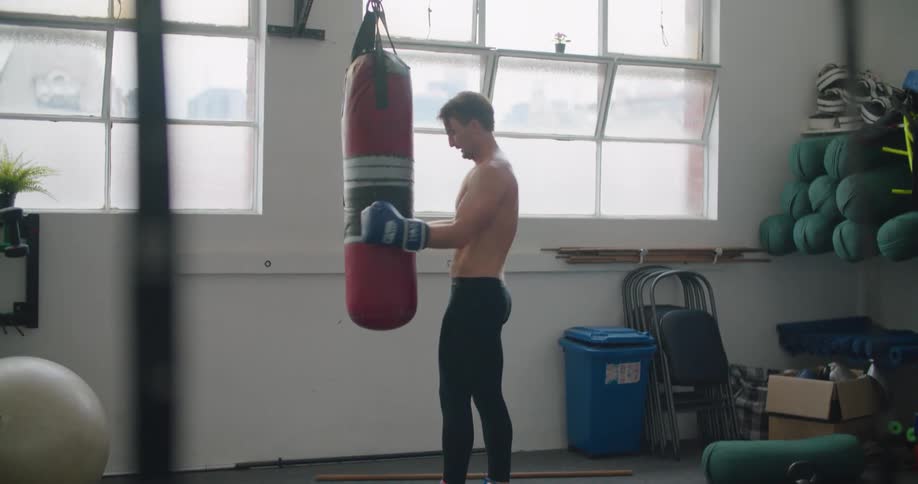 A man with boxing gloves next to a heavy bag with a wall and windows in the background