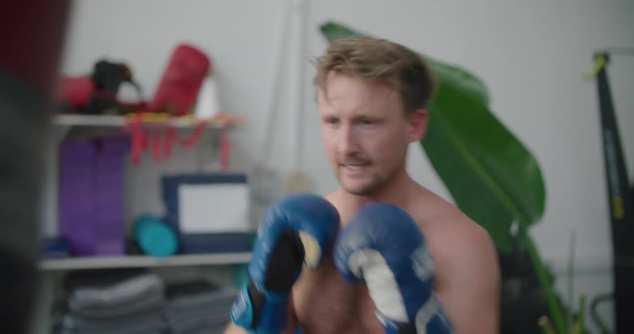 Shirtless muscular man practices boxing with a punching bag in a gym