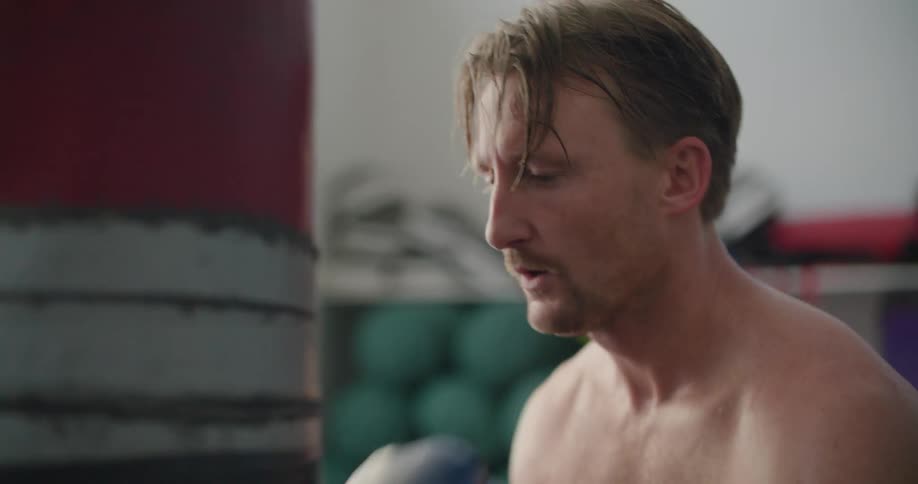 A man resting his head against a boxing bag stands back up straight and begins punching the bag