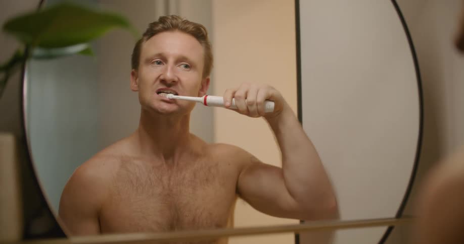 A man stands in front of a mirror and brushes his teeth and then looks at them