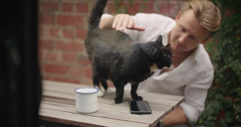 A man pets and kisses a cat while sitting at a picnic table outdoors during the day