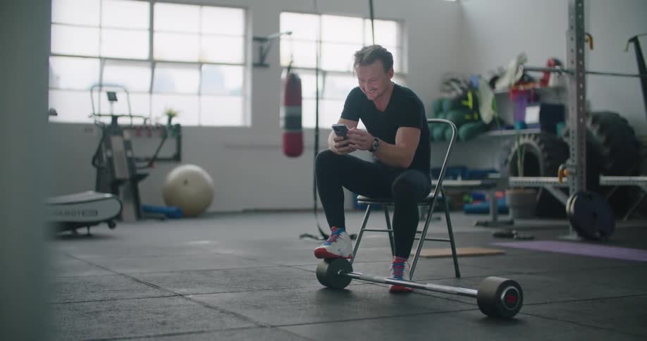 A man sits on a chair in a workout room with his foot resting on a barbell and scrolls on his cell phone