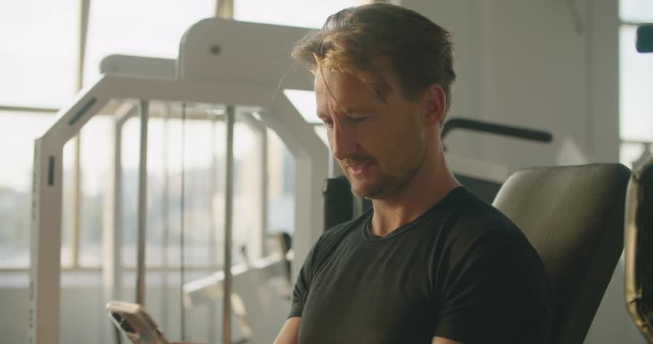 A man scrolls through his smartphone while sitting on equipment at the gym