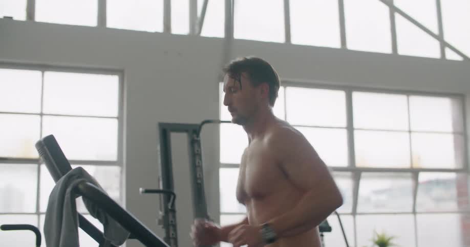 A man in a gym running on a treadmill with equipment and windows in the background
