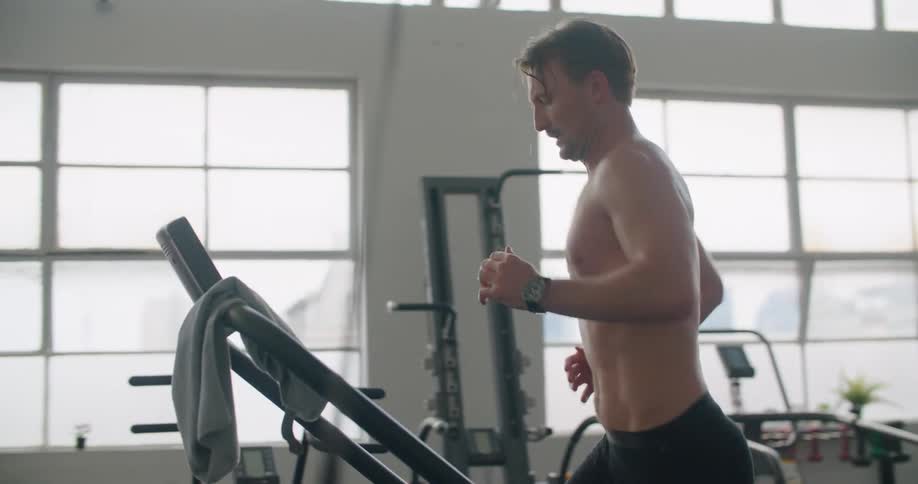 A man in a gym running on a treadmill with equipment and windows in the background