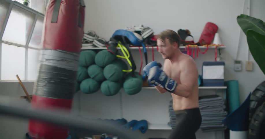 A shirtless man strikes a punching bag in a gym during the day