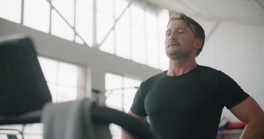 A man in a gym running on a treadmill with equipment and windows in the background
