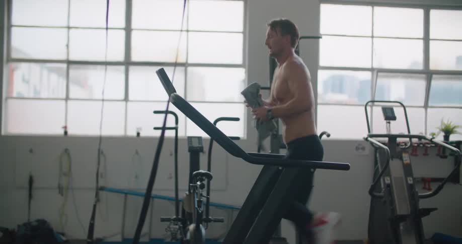A man in a gym running on a treadmill with equipment and windows in the background