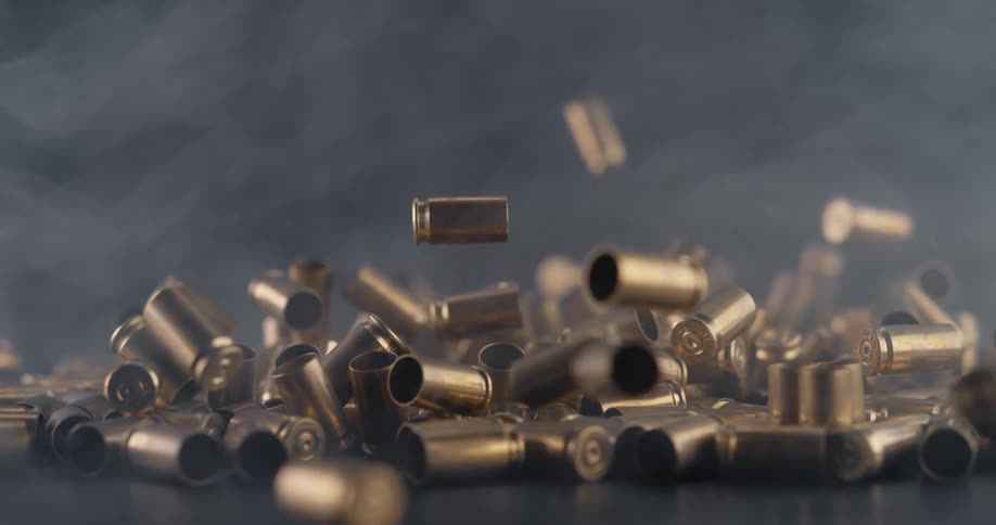A large number of spent bullet casings fall down from above and land in a pile as smoke drifts through the air
