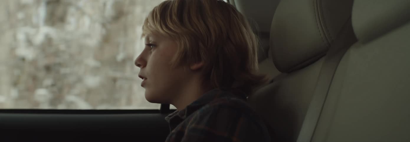 Blonde boy riding in car leans to look out window at snow covered trees and houses