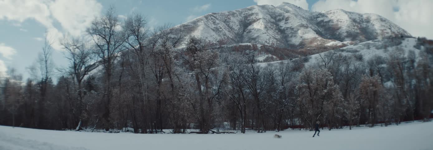 A dog follows a child running through the snow with trees and a snow covered mountain in the background