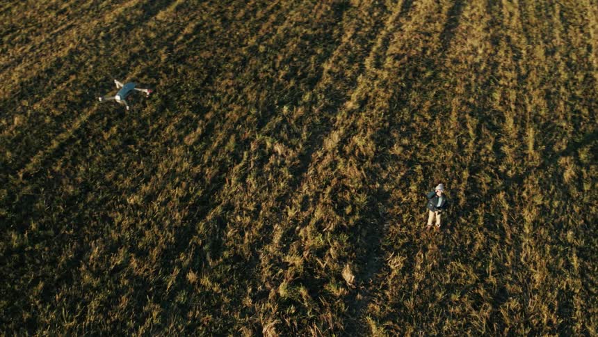 A person with a hat and jacket flying a drone on a grassy field