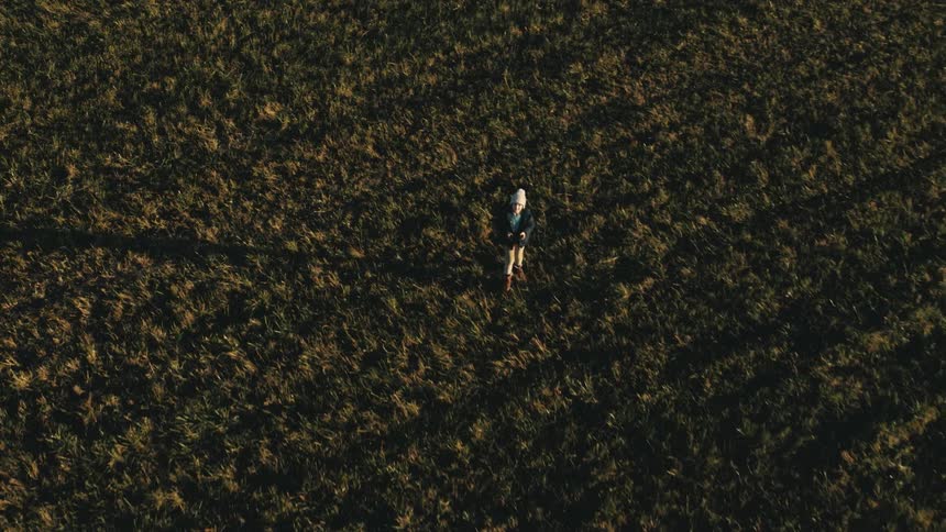A boy walks through a vast field and looks up while operating a drone