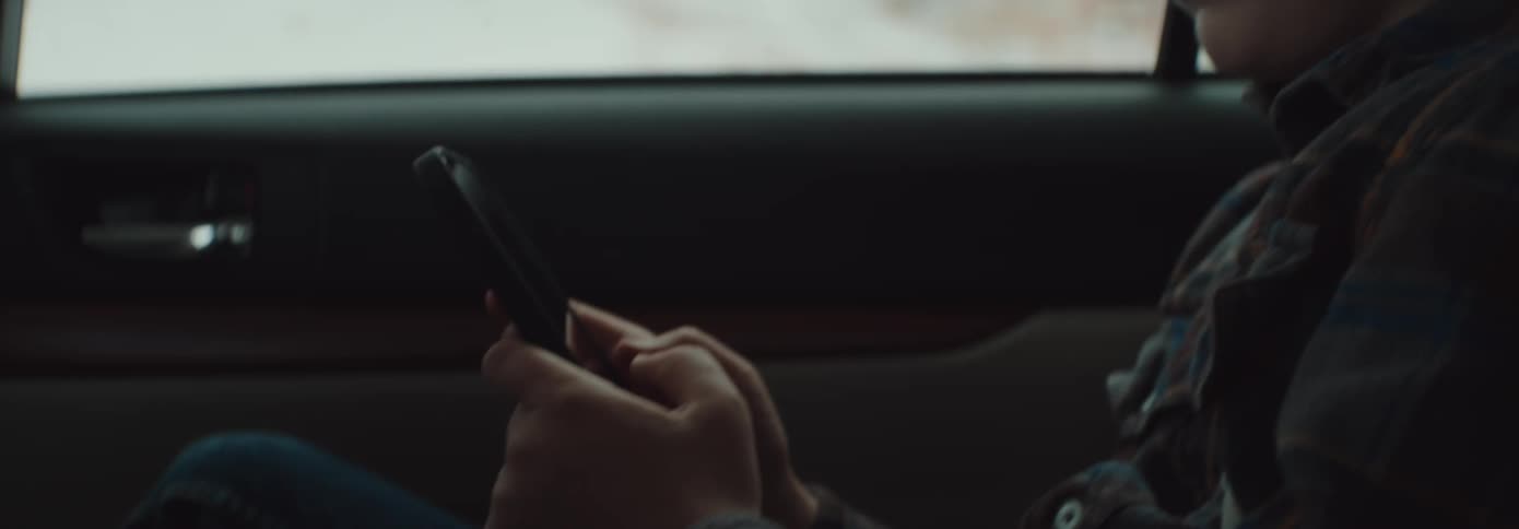 A boy sitting in a vehicle playing with a smart phone with trees and snow in the background