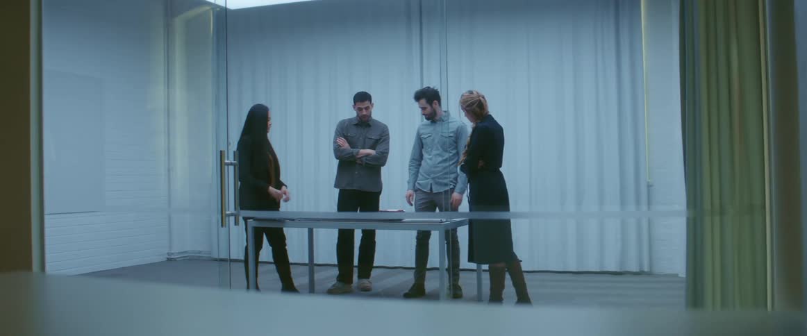 A group of coworkers speak while gesturing at objects on a table in an office during the day