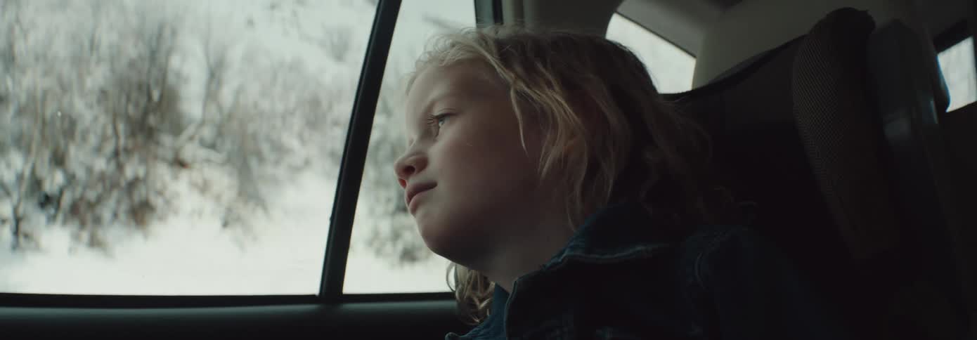 A child gazes out of the window while riding in the backseat of a vehicle moving through a snowy countryside during the day