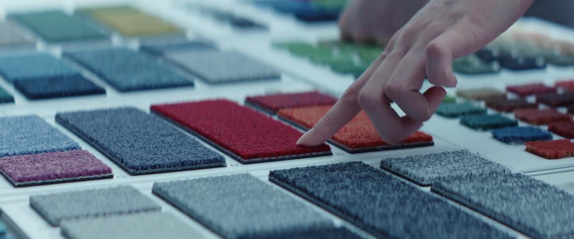A finger pointing and touching the surfaces of different colored small rug samples