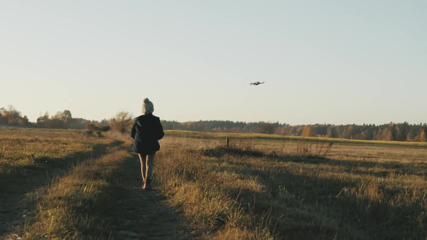 Child walks along partly overgrown dirt road past fields toward forest on horizon guiding drone