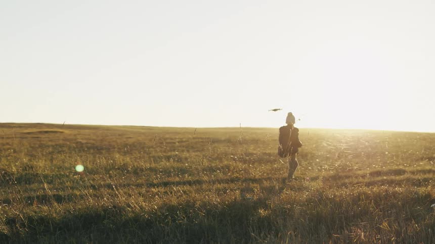 Child wearing a knit hat and a jacket is flying a drone in an open field with fall colored trees in the distance
