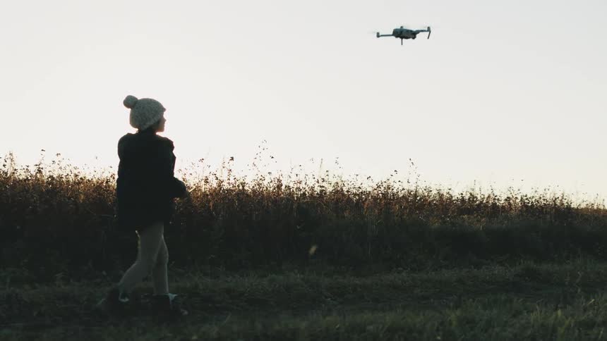 Young person walks past tall vegetation in field following drone side lit by sun