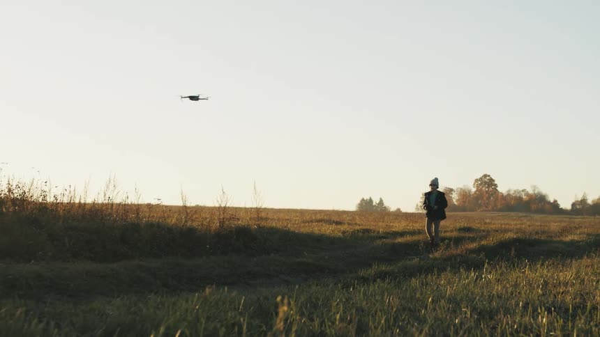Child wearing a knit hat and a jacket is flying a drone in an open field with fall colored trees in the distance