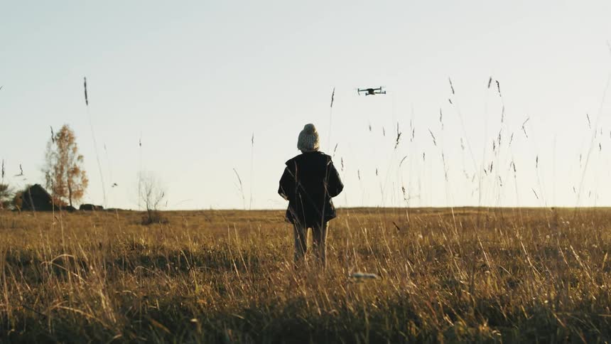 A girl with a hat and jacket flying a drone on a grassy field with the sky and clouds in the background