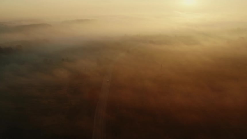 An aerial view of clouds covering a grassland with a car driving down a road