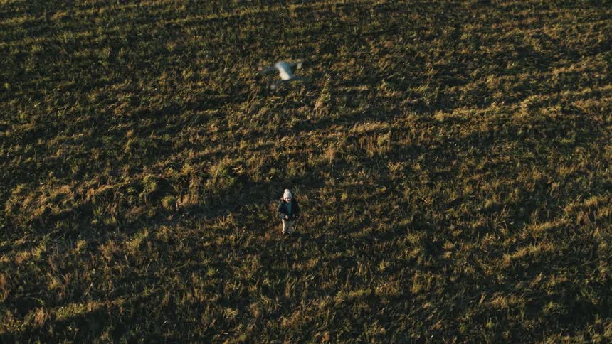 A person with a hat and jacket flying a drone on a grassy field