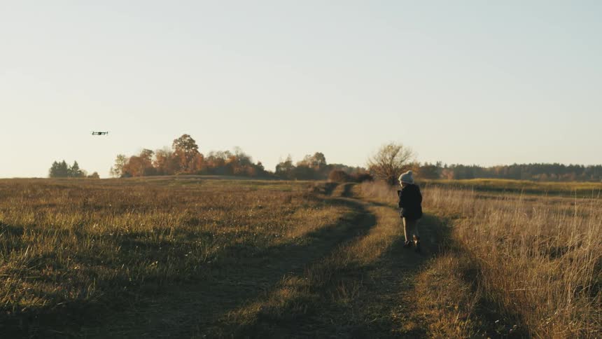 Boy wearing white wool hat and black coat flies a drone in the field